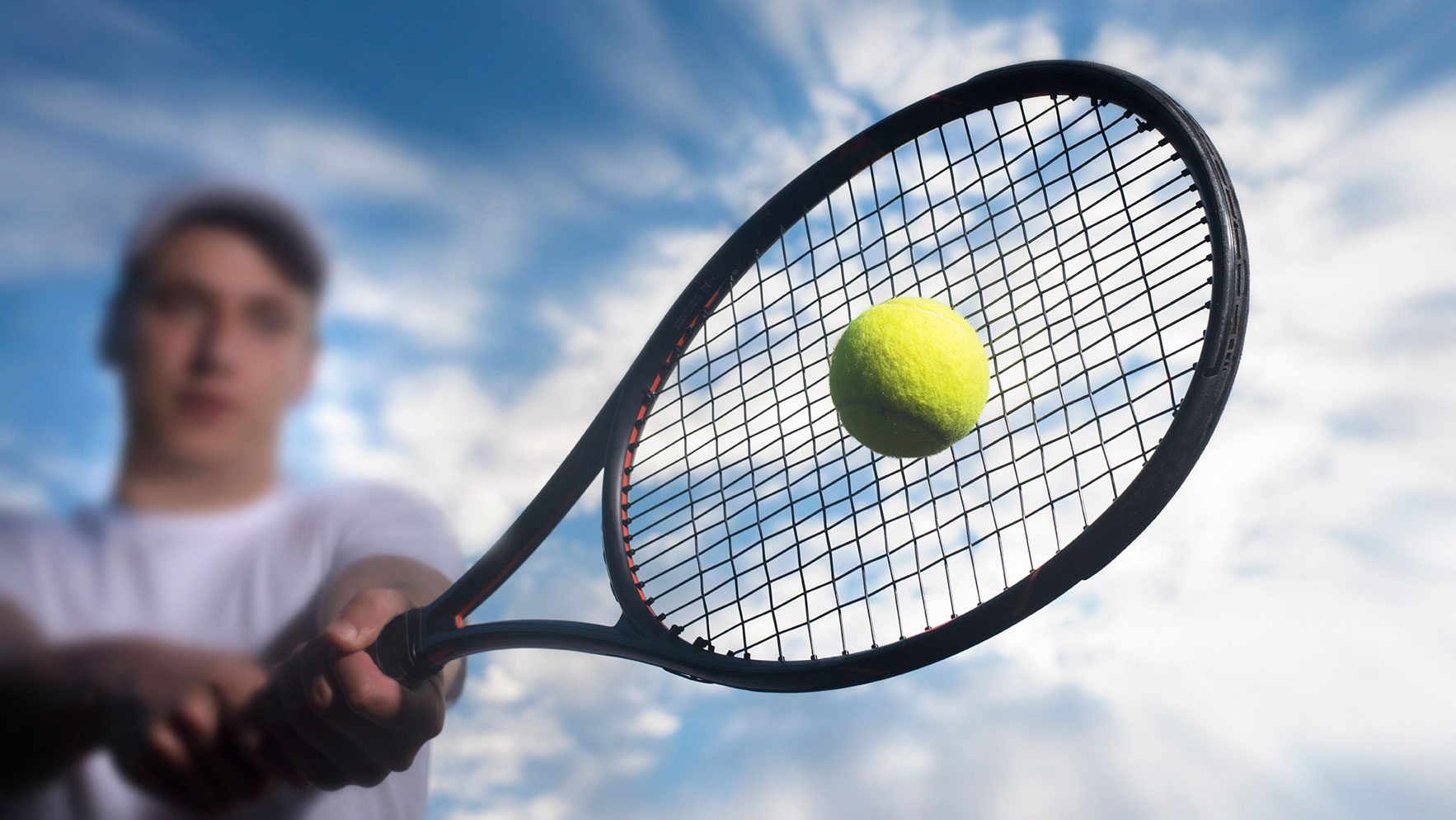 Close up image of a tennis racket hitting a tennis ball. A man is swinging the racket. Blue sky and clouds in the background.