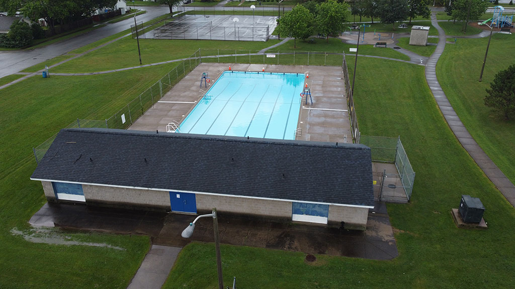Drone overhead image of the pool building and swimming pool. It shows the basketball court, splashpad, walking path and playground in the background.