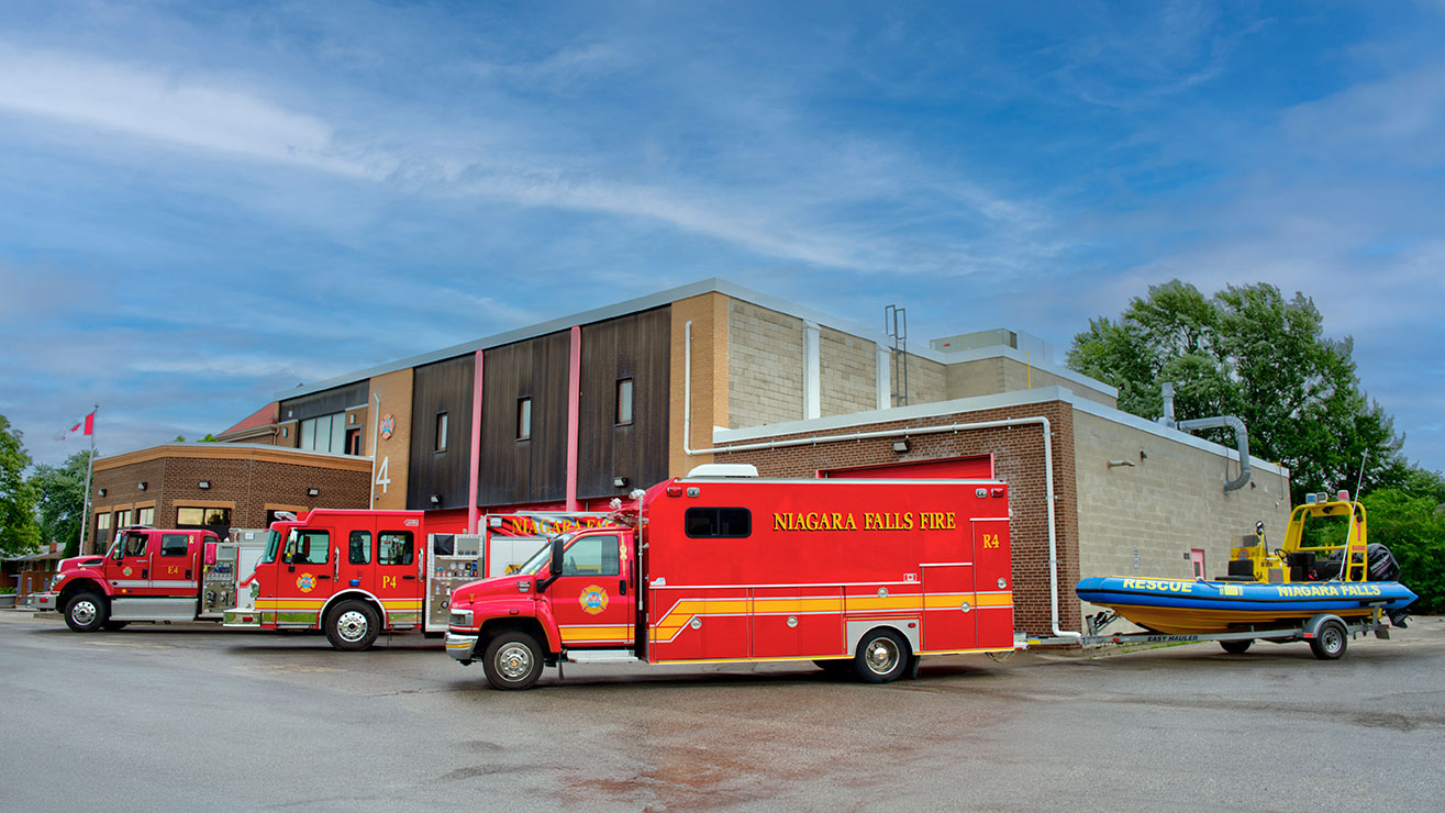 Photo of Fire Station 4. It shows three fire trucks parked in front with one pulling a rescue boat.  The building is in the background with a blue sky above.