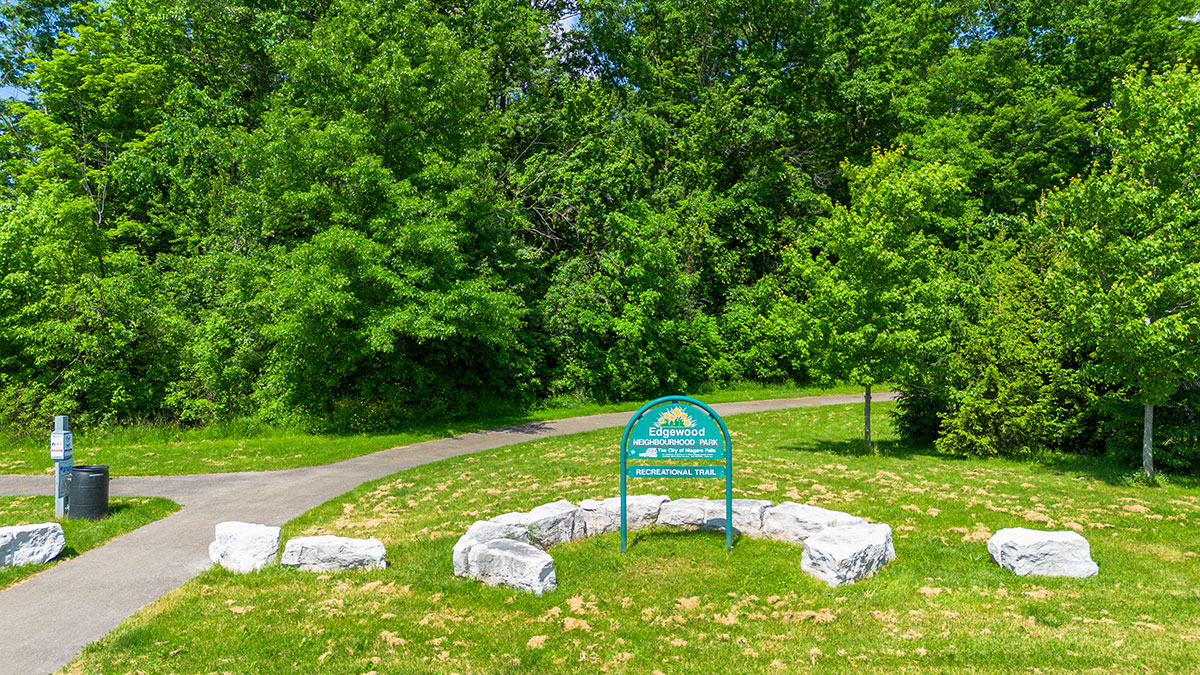 Photo of the Edgewood Woodlot Park Sign. It is surrounded by green grass and landscape rocks. There is a pathway to the left of the sign that goes through green trees.