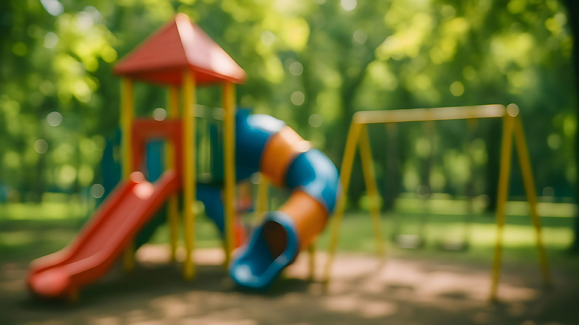 Blurry image of a playground and swing set. There are lots of green trees in the background.