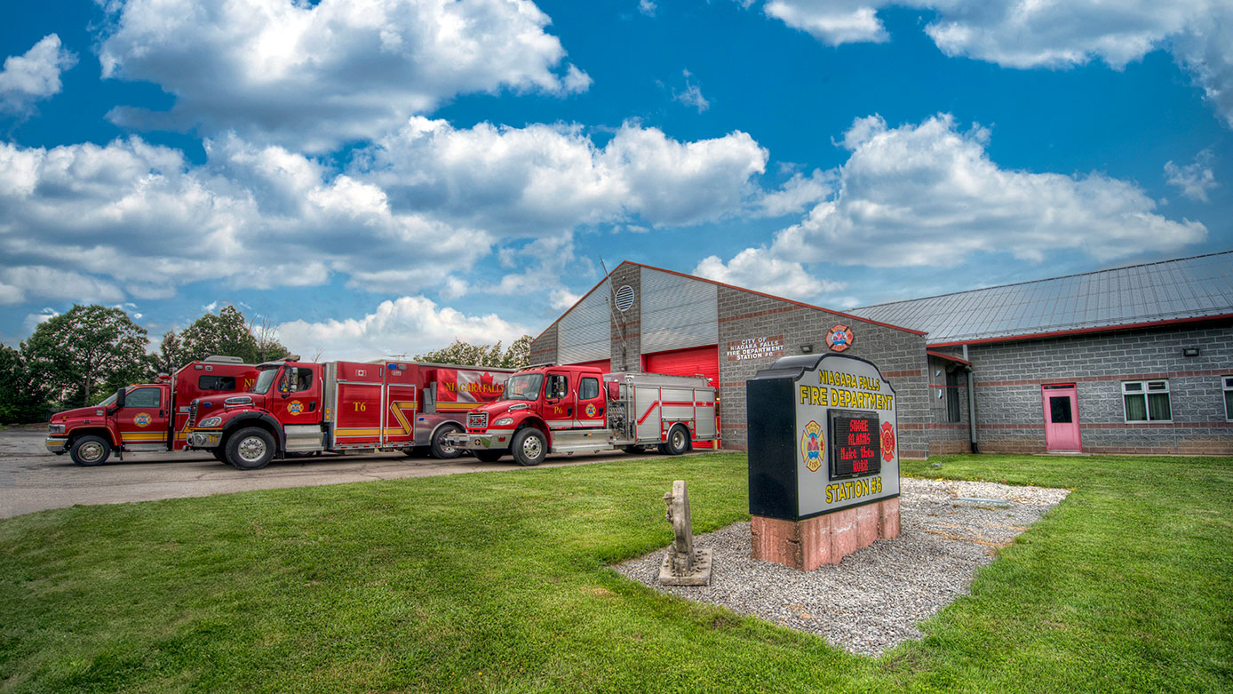 Photo of Fire Station 6.  Fire trucks are parked in front. There is a digital message sign in the middle of the green grass.  A blue sky is above.