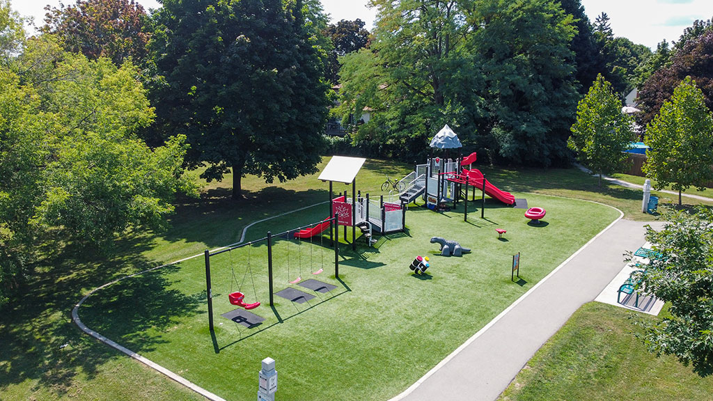Drone overhead image of the Royal Manor Park playground in red colours. Trees surround the park and there are park benches to the right next to the walking path.