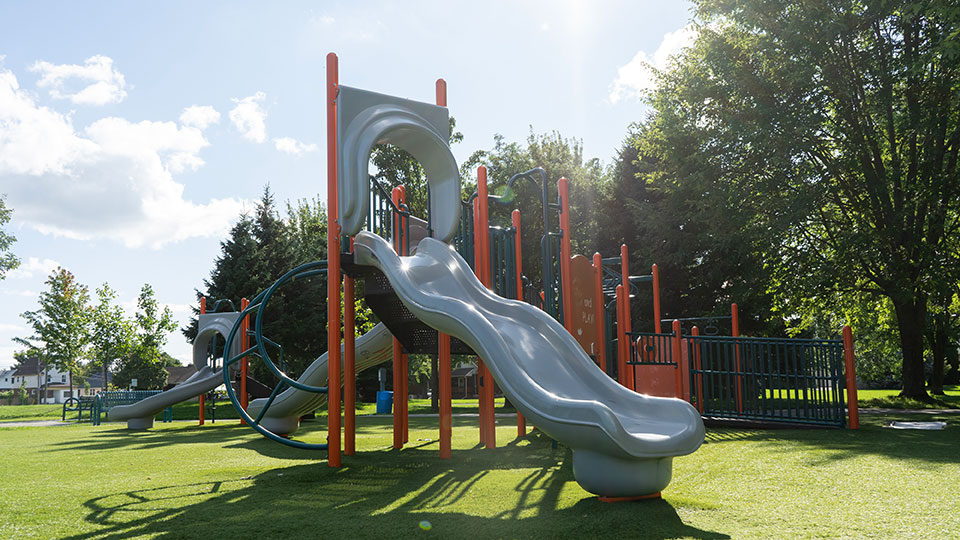 Maple Street Park slide and playground on green turf. Sunny day with green trees in the background. 