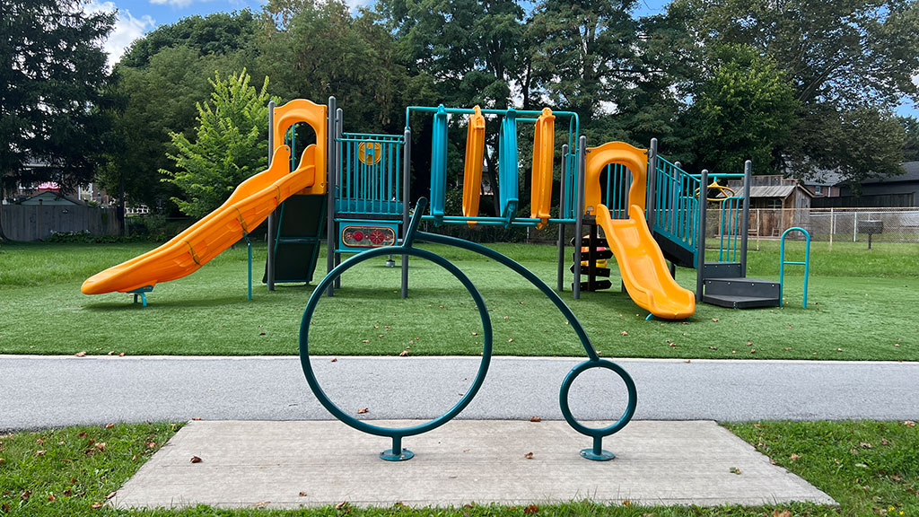 Playground and slides at Alexander Park on a green turf with a bike shaped bicycle rack in the foreground.
