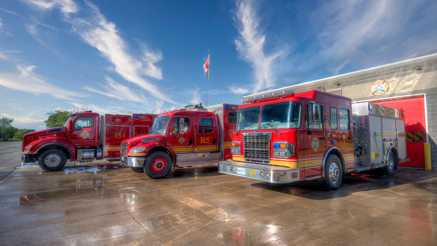 Image of Fire Station 5. Three fire trucks are parked in front.  There is a Canada flag on a pole in the background and blue sky above.